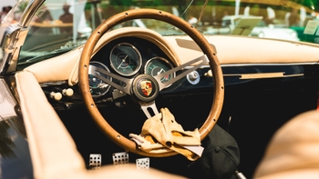 Interior de un Porsche clásico con volante de madera, velocímetro y guantes de piel, mostrando el emblema de Porsche en el centro.