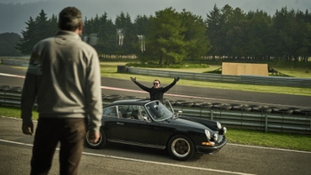 Un hombre se asoma por el techo de un Porsche 911 clásico de color negro, extendiendo los brazos con entusiasmo, mientras el coche está parado en el borde de una pista de carreras. Otro hombre, visto de espaldas, se encuentra frente al coche observando la escena. El fondo muestra árboles y un día nublado.