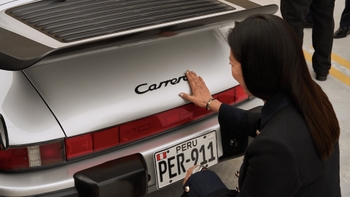Una mujer con cabello oscuro y chaqueta azul toca suavemente el emblema "Carrera" en la parte trasera de un Porsche 911 de color plata. El coche tiene un alerón trasero negro tipo "cola de ballena" .