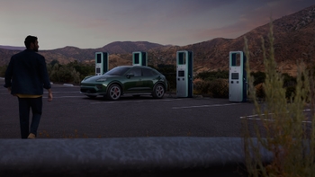 Hombre caminando hacia un Porsche Macan eléctrico verde cargando en una estación, con montañas al atardecer de fondo.