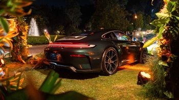 Vista trasera de un Porsche verde oscuro en un entorno elegante con iluminación nocturna, rodeado de vegetación y con una fuente en el fondo.