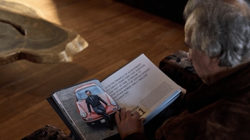 Un hombre, visto desde atrás, está sentado leyendo un libro de gran formato sobre Porsche. En la página izquierda del libro, se muestra a un hombre sentado elegantemente sobre un Porsche 911 clásico de color rojo. La página derecha contiene texto y el título "Porsche 911", con una iluminación cálida que resalta los detalles del mobiliario de madera.