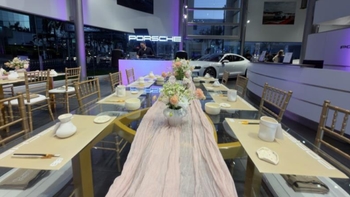 Toma a lo largo de la mesa decorada, de fondo se ve el logo de Porsche luminoso y un Porsche Taycan color blanco.