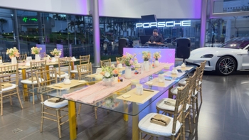 Angulo de todas las mesas decoradas vacias, buena ambientación e iluminacion, de fondo el logo luminoso de Porsche y un Porsche Taycan color blanco.