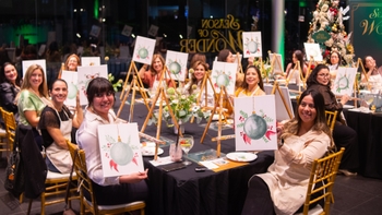 Grupo de mujeres mostrando sus pinturas en el evento "Season of Wonder" de Porsche, con ambiente navideño y lienzos con motivos festivos.