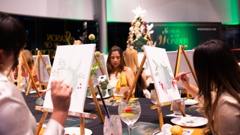 Dos mujeres sonrientes posando en un evento "Season of Wonder" de Porsche, con lienzos pintados y un ambiente festivo.