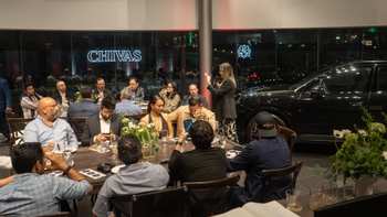 Grupo de personas reunidas en una cena de evento con la marca Chivas visible en el fondo y un Porsche negro exhibido al lado.