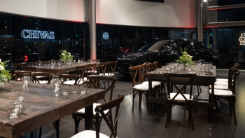 Concesionario Porsche preparado para un evento con mesas rústicas, flores y un Porsche negro en exhibición bajo la luz nocturna de la ciudad.
