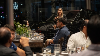 Mujer dando un discurso en un evento Porsche, con un Porsche Cayenne negro de fondo y asistentes sentados alrededor de una mesa.