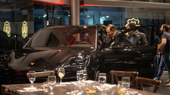 Mujer dando un discurso en un evento Porsche, con un Porsche Cayenne negro de fondo y asistentes sentados alrededor de una mesa.
