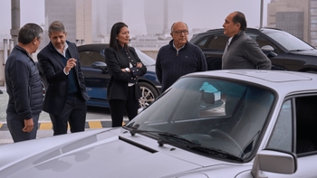Un grupo de cinco personas (cuatro hombres y una mujer) se encuentra de pie en lo que parece ser una terraza o azotea, con edificios urbanos al fondo. En primer plano, se observa parte del capó y el parabrisas de un Porsche 911 de color plata. Las personas están conversando, algunas señalando.