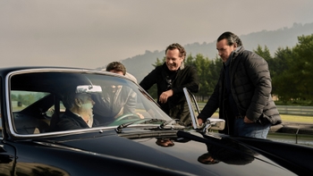 Un grupo de cuatro hombres interactúa alrededor de un Porsche 911 clásico de color negro. Uno de ellos está sentado al volante del coche, mientras los otros tres se inclinan y conversan con él desde fuera, mostrando sonrisas y gestos amigables. El entorno sugiere un exterior con colinas al fondo.