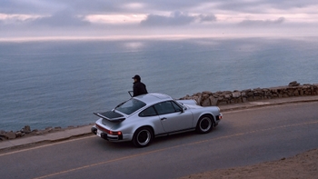 Un Porsche 911 Carrera clásico de color plata está estacionado en una carretera sinuosa junto a un acantilado. Una persona, vestida con chaqueta oscura y gorra, se apoya en el coche mirando el vasto océano bajo un cielo nublado, creando una atmósfera contemplativa.