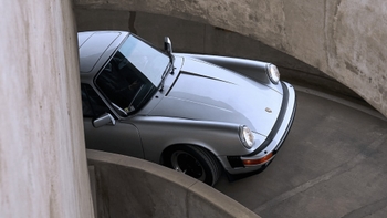 Un Porsche 911 clásico de color plata brillante asciende por una rampa espiral de concreto de varias plantas. La imagen se toma desde una perspectiva elevada, destacando las formas curvas de la estructura de hormigón que rodea el vehículo.