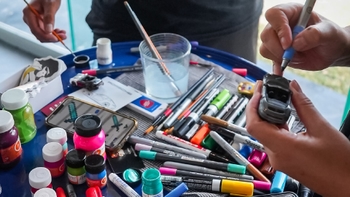 Manos de personas personalizando coches de juguete con marcadores y pinceles. Varios materiales de arte en una mesa.