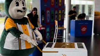 Una mascota sonriente con vestimenta blanca y verde en un evento, con personas y banners de Porsche al fondo.