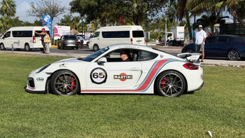 Porsche GT4 blanco con livery Martini y número 65 aparcado sobre el césped con espectadores al fondo.