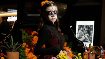 Niña con maquillaje de Catrina y flores de cempasúchil, posando frente a la cajuela de un Porsche decorada para el Día de Muertos.