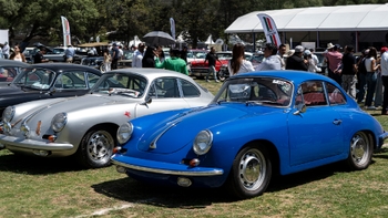 Dos Porsche 356 clásicos, uno azul y otro plata, estacionados sobre césped en un evento automovilístico con personas y carpas de fondo.