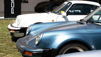 Primer plano de dos Porsche 911 clásicos, uno azul y otro blanco, con un cartel "PORSCHE" de fondo en un evento.