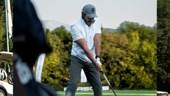 Golfista en camisa blanca y gorra gris se prepara para un golpe en el campo de golf, con logo Porsche sutil al fondo y árboles.