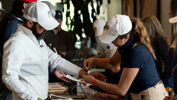 Participantes de evento de golf siendo registrados, una mujer coloca un brazalete a un hombre con gorra y barba en la mesa.