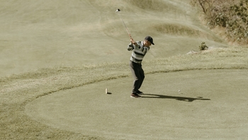 Golfista golpeando la bola en un campo de golf con terreno ondulado.