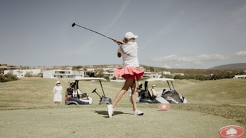 Golfista femenina realizando un swing en el campo de golf, con carritos de golf y paisaje de fondo, en la Porsche Golf Cup.
