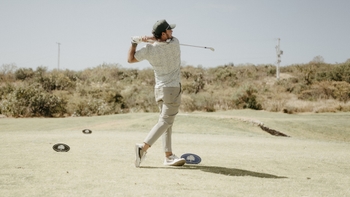 Golfista realizando un swing en un campo de golf soleado, con vegetación al fondo.