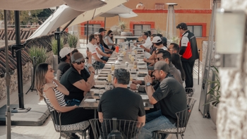 Grupo de personas disfrutando de una comida al aire libre en una terraza con sombrillas y vegetación, con un camarero vestido con el logo de Porsche.