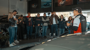 Grupo de personas escuchando una presentación en un concesionario Porsche moderno, con varios autos exhibidos en el fondo.