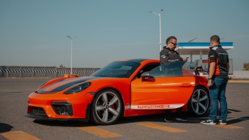 Porsche 718 Cayman GT4 RS naranja con detalles en negro, dos personas junto al auto en un ambiente soleado.