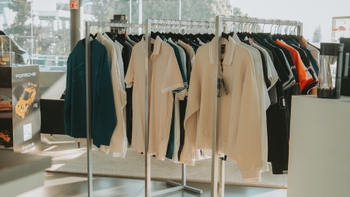 Estantes con ropa de vestir casual y deportiva, incluyendo polos y sudaderas, dentro de un concesionario Porsche, con un expositor del coche de juguete Porsche de fondo.