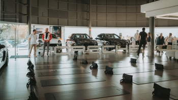 Amplia vista de un evento en un concesionario Porsche, donde se observan numerosas esterillas de yoga preparadas en el suelo y varios coches de lujo en exhibición. Diversas personas se encuentran en el lugar interactuando.
