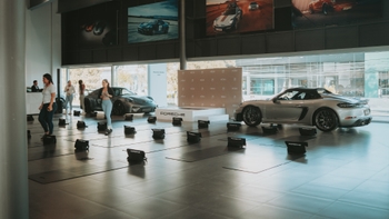 Un evento de yoga se lleva a cabo en el interior de un concesionario Porsche, con esterillas alineadas, participantes en ropa deportiva y dos coches Porsche de lujo en exhibición.