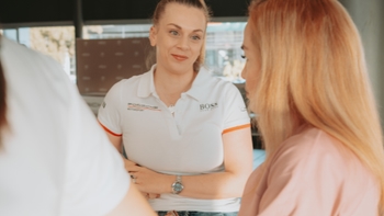 Una mujer con un polo blanco de Porsche Motorsport y Boss, conversando atentamente con otra persona en un entorno de evento.