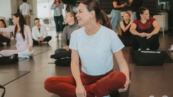 Mujer sonriente sentada en esterilla de yoga durante un evento en concesionario Porsche, con otras participantes en segundo plano.