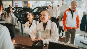Dos mujeres sonriendo y conversando en un mostrador dentro de un concesionario Porsche, con un hombre en ropa deportiva en el fondo.