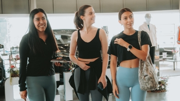 Tres mujeres en ropa deportiva sonriendo y conversando en el área de exhibición de un concesionario Porsche, con un Macan oscuro de fondo.