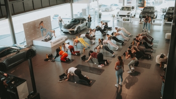 Grupo de mujeres haciendo yoga o pilates (plancha) en un concesionario Porsche con varios modelos: 718 Cayman GT4 RS, Cayenne y Macan.