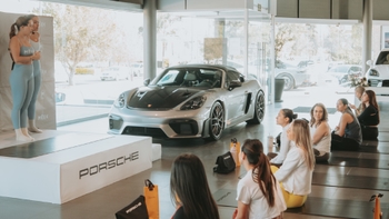 Mujeres participantes en una clase de yoga o evento, sentadas frente a instructores y un Porsche 718 Cayman GT4 RS.