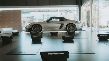 Primer plano de un coche Porsche 718 Spyder RS plateado en exhibición dentro de un concesionario, rodeado de esterillas de yoga con el logo de Porsche y el fondo con el branding de MEEK.