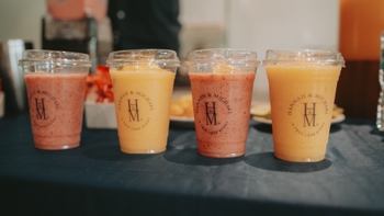 Cuatro vasos de smoothies de frutas con el logo "Hannah & Michael" en una mesa oscura, listos para servir.