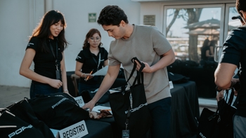 Un hombre recogiendo una bolsa de bienvenida con logo Porsche en una mesa de registro, atendida por dos mujeres del staff de Porsche, en un evento.