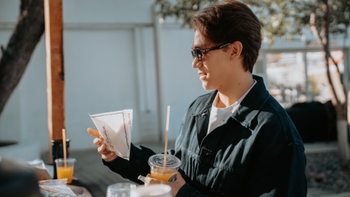 Un joven sonriente con gafas de sol, sosteniendo un zumo de naranja y una pequeña bolsa de papel de "La Panoteca", disfrutando de un refrigerio al aire libre.