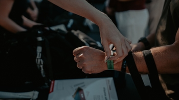 Close-up de unas manos colocando una pulsera de acceso verde en la muñeca de una persona en un evento Porsche, con un fondo oscuro y un objeto blanco parcialmente visible.
