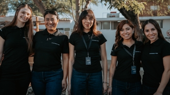 Un grupo de cinco mujeres sonrientes, vestidas con uniformes negros de staff de Porsche, posan para la cámara en un evento al aire libre.