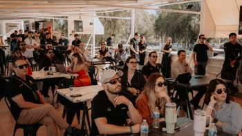 Grupo de personas escuchando en un evento, sentados y de pie bajo una carpa, con mesas y bebidas.