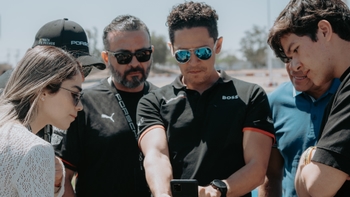 Un grupo de personas, con vestimenta de Porsche y gorras "Track & Friends", se congrega para ver algo en un smartphone.