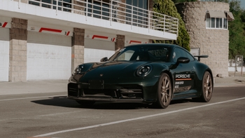 Un elegante Porsche 911 verde oscuro con la marca "Porsche Driving Center" en su lateral, estacionado en la pista frente a los garajes de boxes en un día soleado.